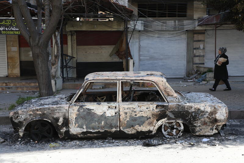 a burned-out vehicle in Jablah town, Lattakia Governorate, following Syria's recent escalation in violence. Access to basic services has been severely disrupted, with some cities still without power.
